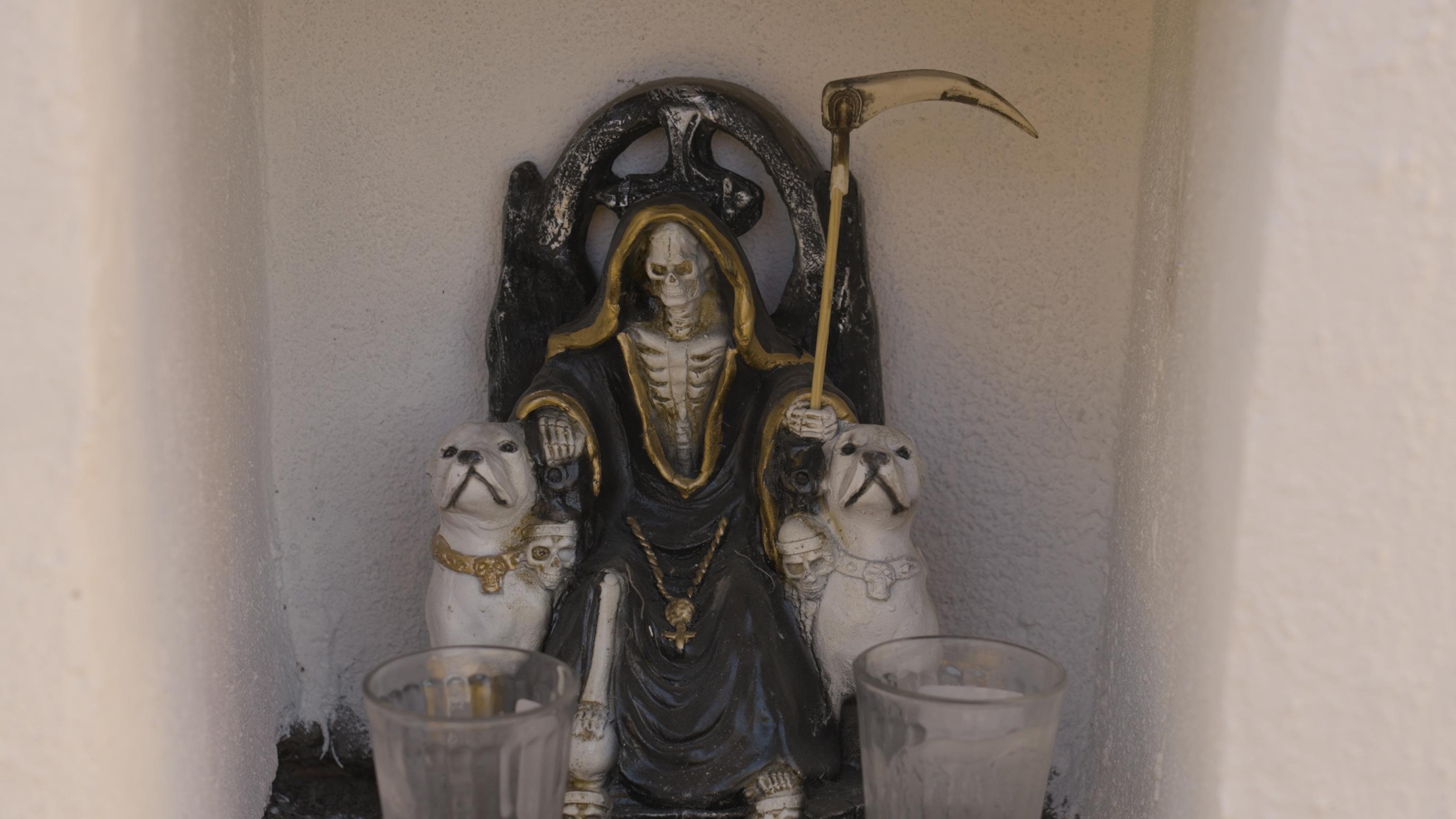 A Santa Muerte shrine where Santa Muerte is seated and has a dog to the left and the right of her chair